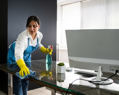 woman janitor with yellow gloves cleaning a glass work desk with cleaning detergent in an office