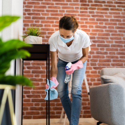 woman janitor with pink gloves holding a cleaning detergent can performing cleaning of a black stand with blue cloth in a home